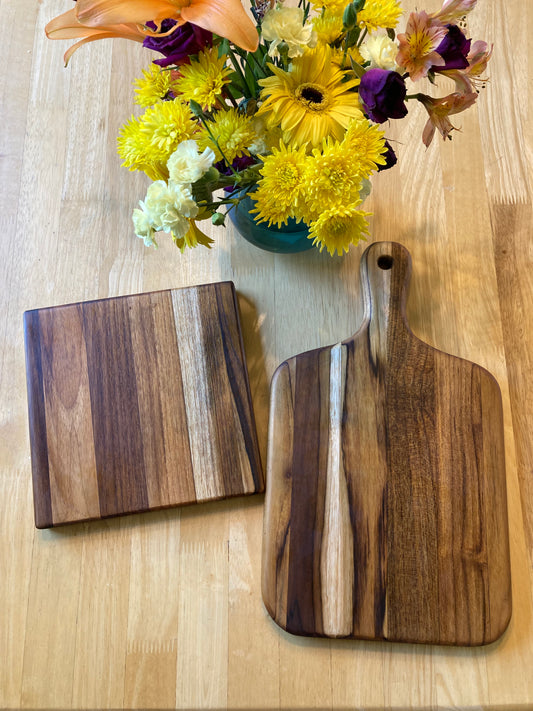 Two wooden teak cutting boards with distinct grain patterns, one larger hand-held paddle board and one smaller square board, displayed on a light wood tabletop next to a yellow flower bouquet.