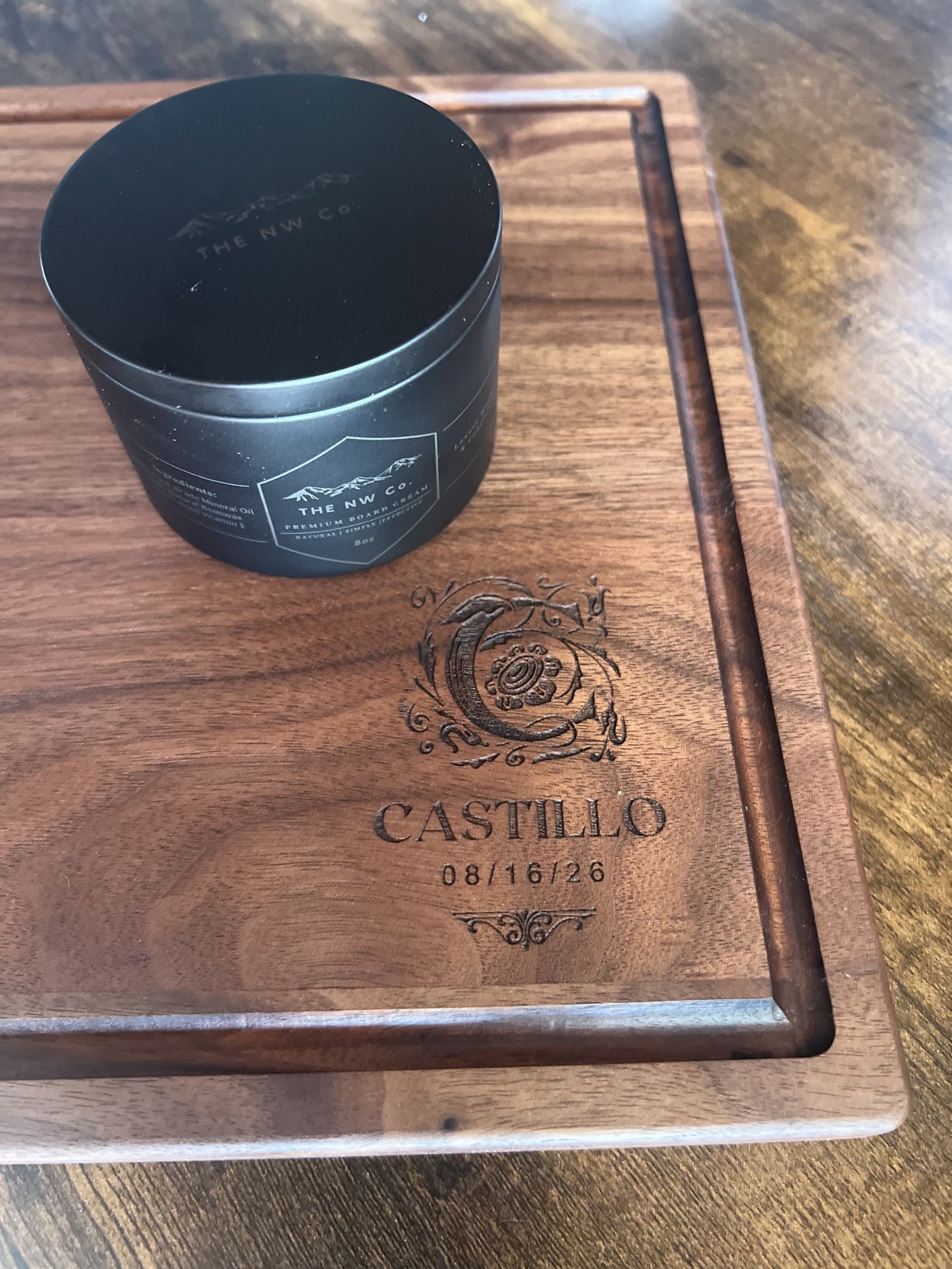 Cutting board engraved with an ornate monogram, family name, and established date, shown with a jar of NW Co. Board Cream