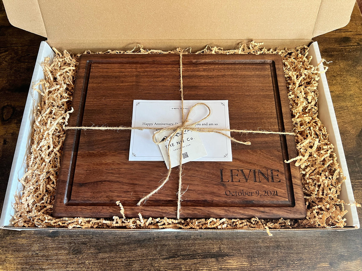 Gift-ready packaging: engraved black walnut cutting board tied with twine and a printed note card in a gift box with kraft crinkle paper