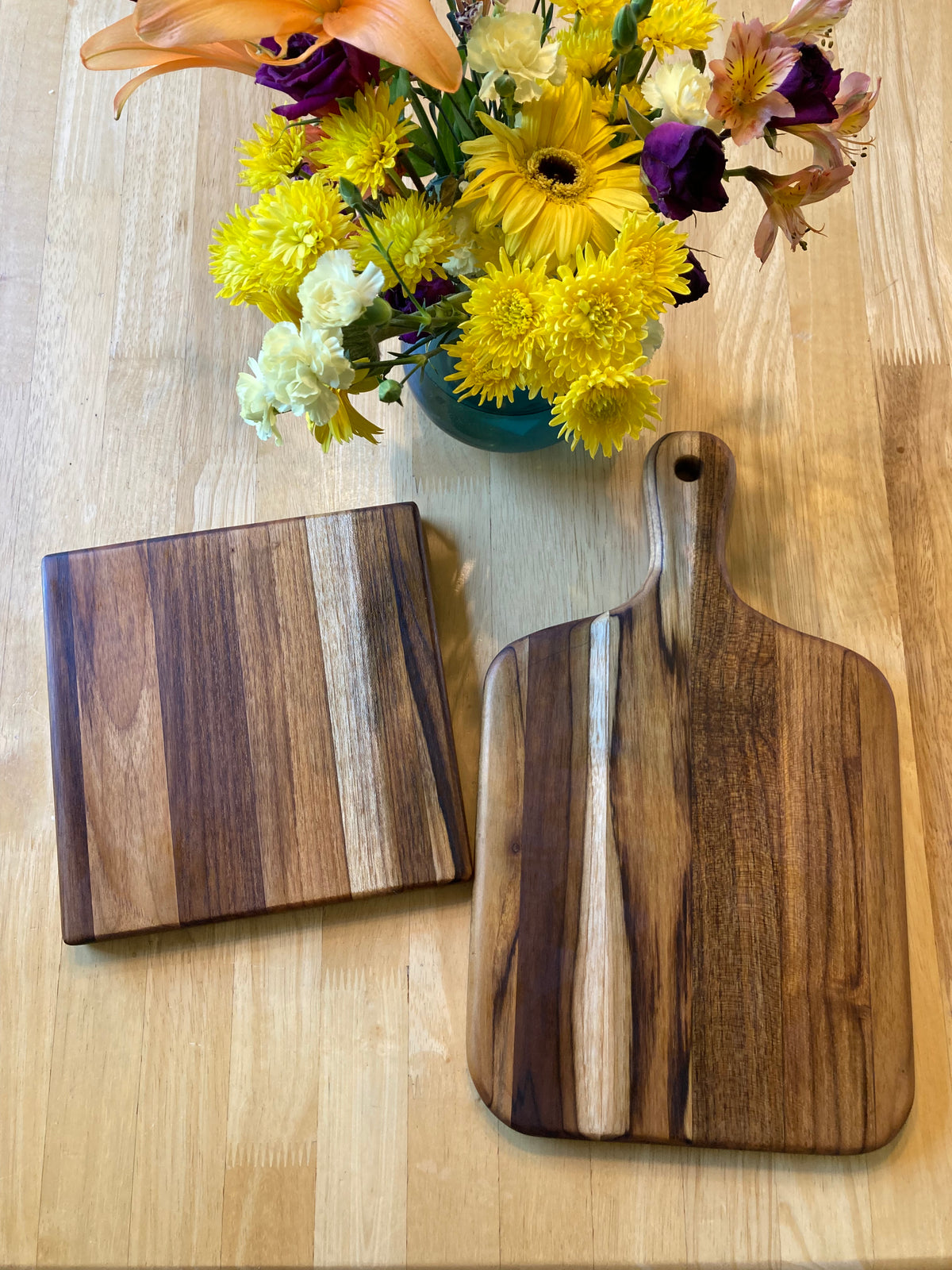 Two wooden teak cutting boards with distinct grain patterns, one larger hand-held paddle board and one smaller square board, displayed on a light wood tabletop next to a yellow flower bouquet.